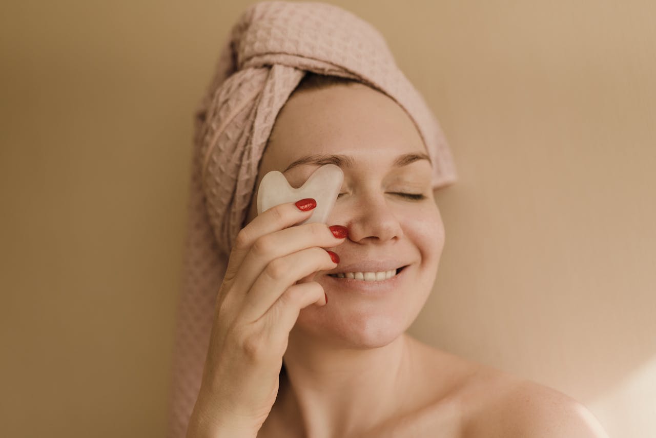 A smiling woman with a towel wrapped around her head using a gua sha tool for a facial massage.